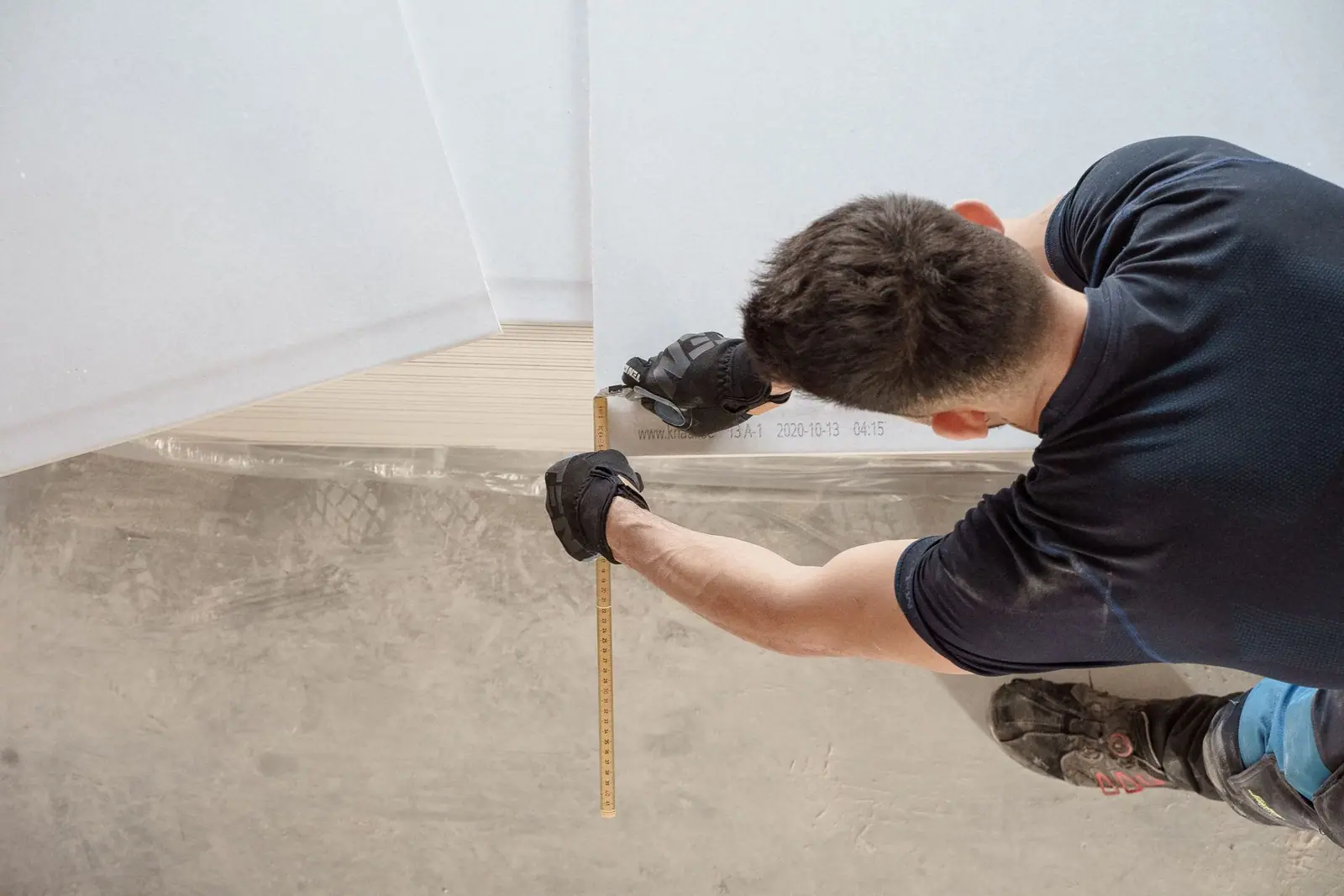 A worker kneeling beside a flooring panel and measuring it with a tape measure