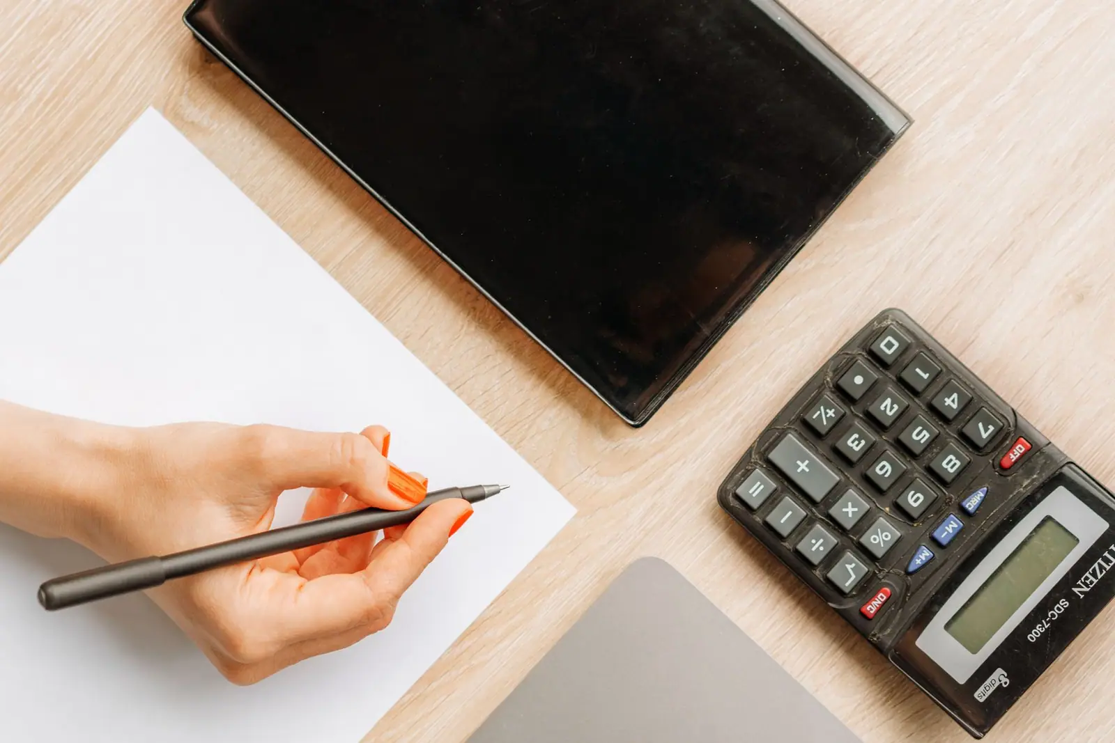 A desk seen from above with a hand writing beside a calculator