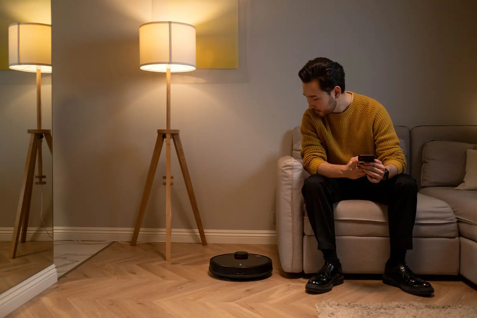 A person holding a phone on a sofa while a black robot vacuum sits on the floor