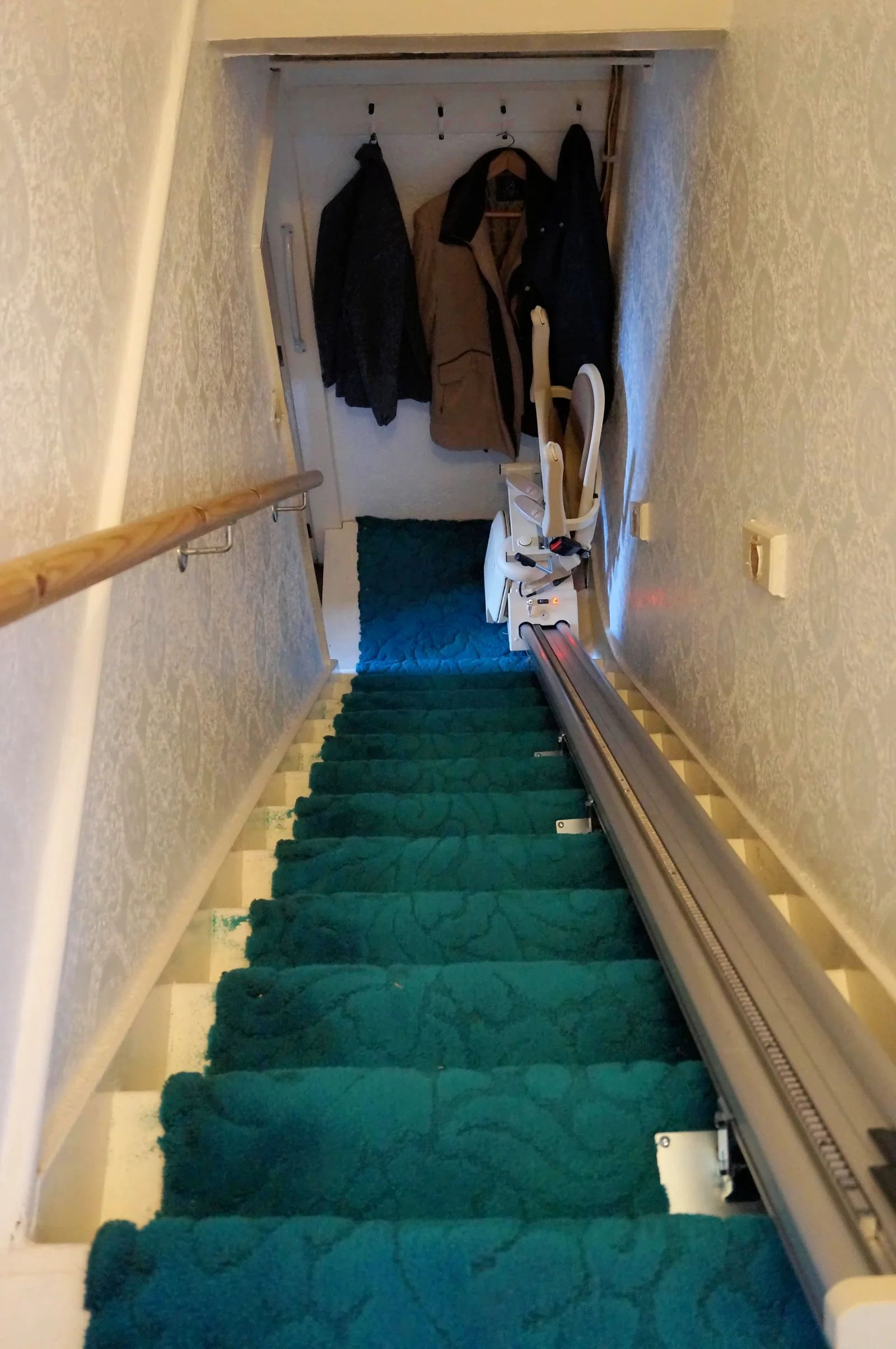 A folded stairlift chair beside a rail on a narrow teal-carpeted staircase