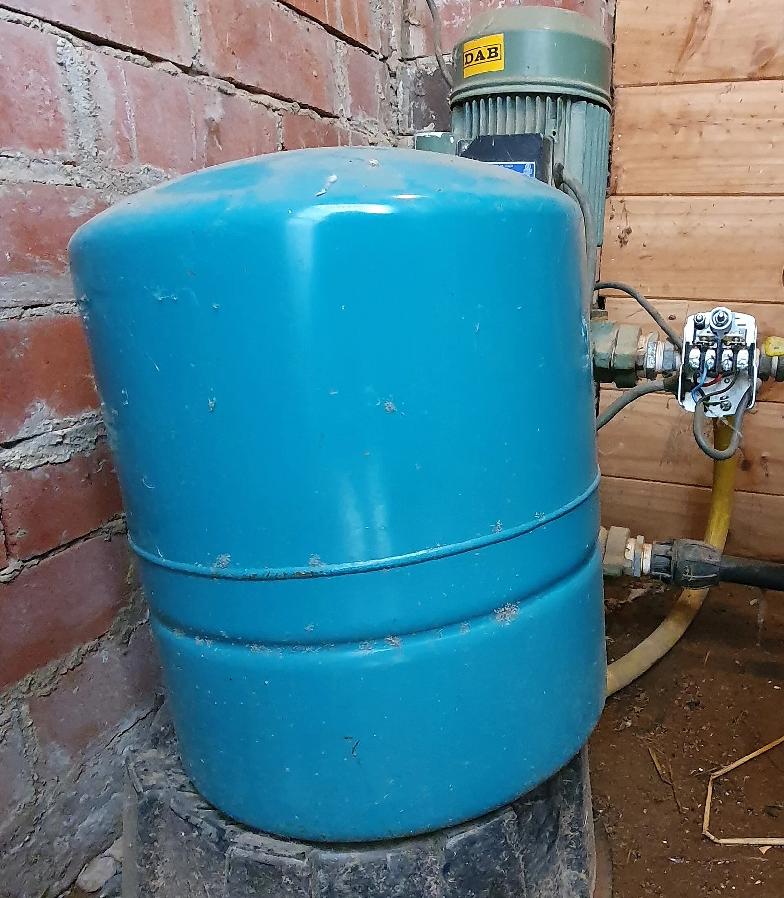 A blue cylindrical pressure tank connected to pipes beside a brick wall